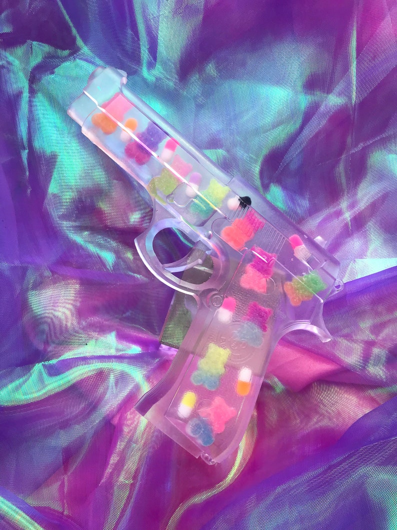 May include: A clear plastic toy gun filled with colorful gummy bears and candy. The gun is lying on a iridescent purple and blue fabric.