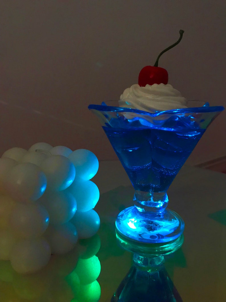 K&ouml;nnte beinhalten: Eine blaue Glassch&uuml;ssel mit einem Topping aus wei&szlig;er Schlagsahne und einer roten Kirsche. Die Sch&uuml;ssel wird von unten beleuchtet und erzeugt einen blauen Schein auf dem Tisch.