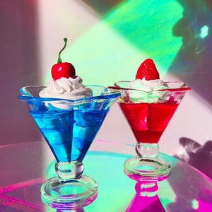 K&ouml;nnte beinhalten: Zwei gl&auml;serne Dessertbecher mit Schlagsahne und einer Kirsche und einer Erdbeere obenauf. Das Dessert im linken Becher ist blau und das Dessert im rechten Becher ist rot.