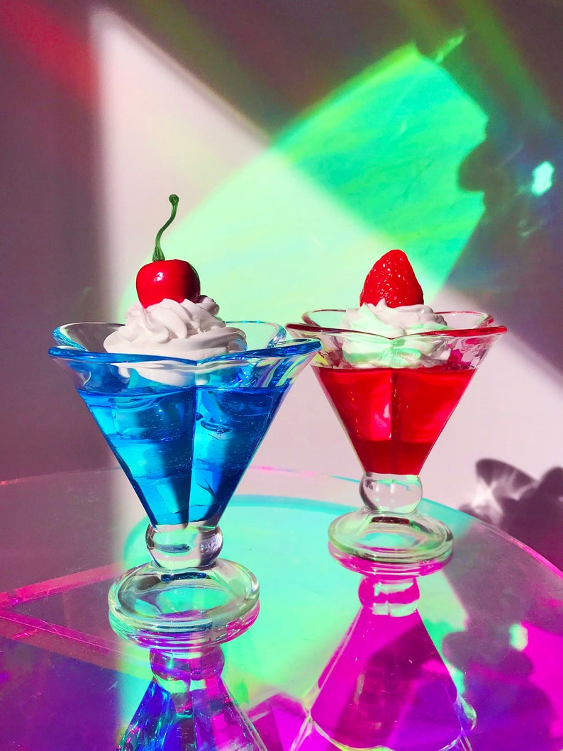 K&ouml;nnte beinhalten: Zwei gl&auml;serne Dessertbecher mit Schlagsahne und einer Kirsche und einer Erdbeere obenauf. Das Dessert im linken Becher ist blau und das Dessert im rechten Becher ist rot.