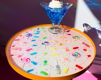 May include: A round, yellow side table with a white top decorated with colourful sprinkles, lollipops, and pills. A blue glass sundae dish with whipped cream and a cherry sits on top of the table.