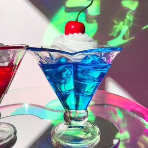 K&ouml;nnte beinhalten: Eine blaue Glasschale in Form einer Eisbecher mit einer wei&szlig;en Schlagsahne-F&uuml;llung und einer roten Kirsche obenauf. Die Schale steht auf einer schillernden Oberfl&auml;che.