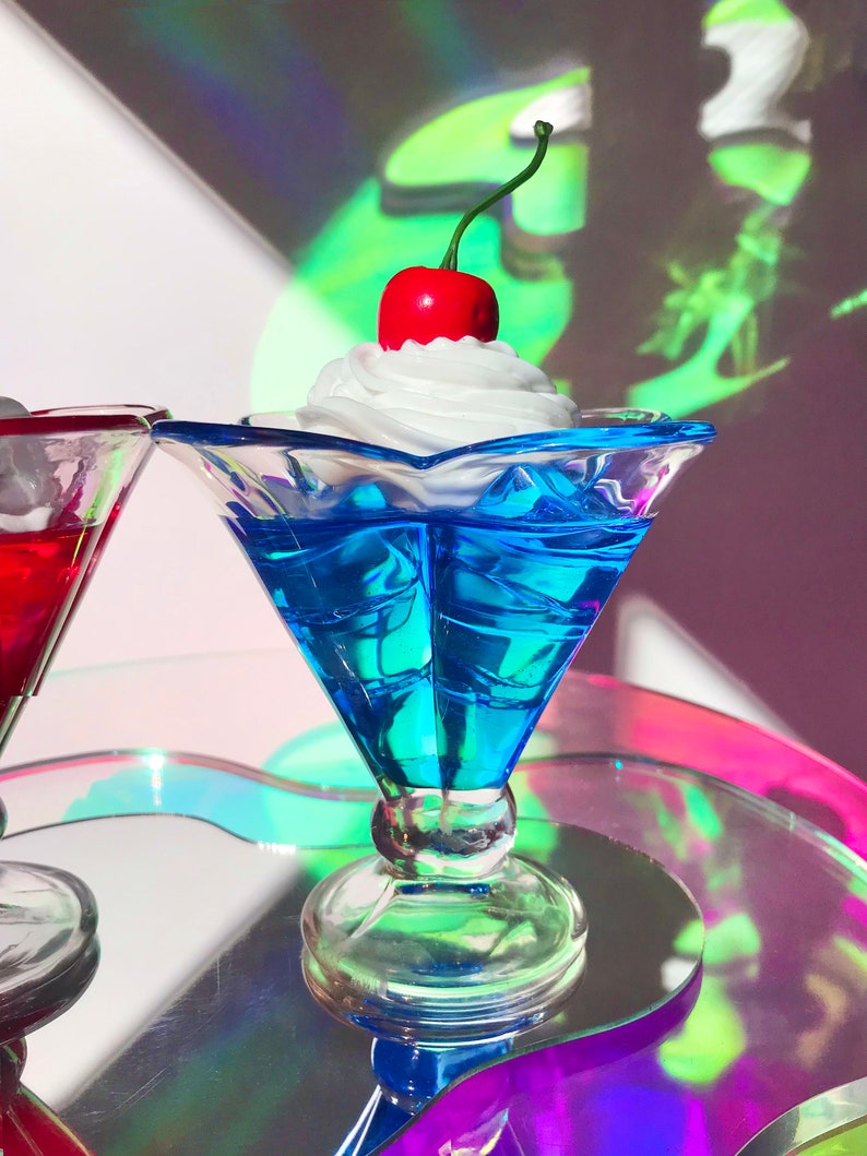 K&ouml;nnte beinhalten: Eine blaue Glasschale in Form einer Eisbecher mit einer wei&szlig;en Schlagsahne-F&uuml;llung und einer roten Kirsche obenauf. Die Schale steht auf einer schillernden Oberfl&auml;che.