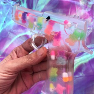 May include: A clear plastic toy gun filled with colorful candy hearts. The gun is held in a hand.
