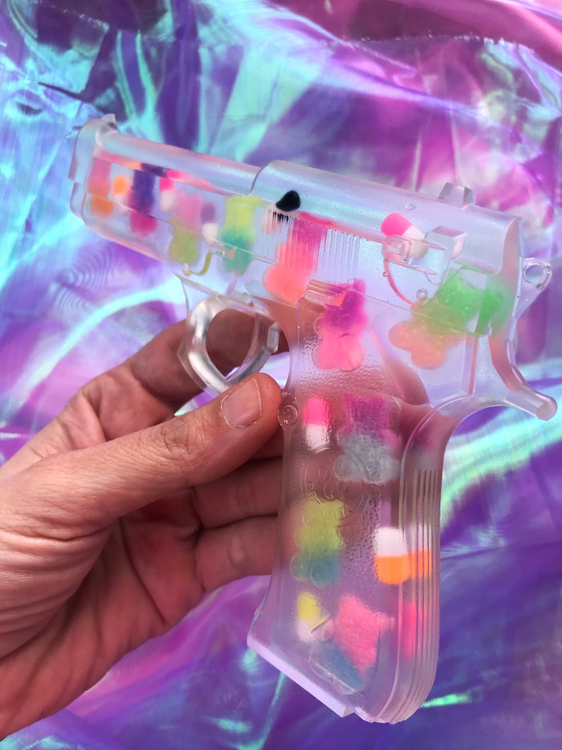 May include: A clear plastic toy gun filled with colorful candy hearts. The gun is held in a hand.