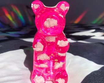 May include: A pink gummy bear figurine with white clouds and glitter inside. The bear is standing on a black and white chequered surface.