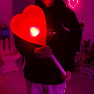 May include: A person holding a large, red, heart-shaped balloon with a white stick. The balloon is lit from within, creating a glowing effect. The background is a pink and purple room with a neon heart sign on the wall.