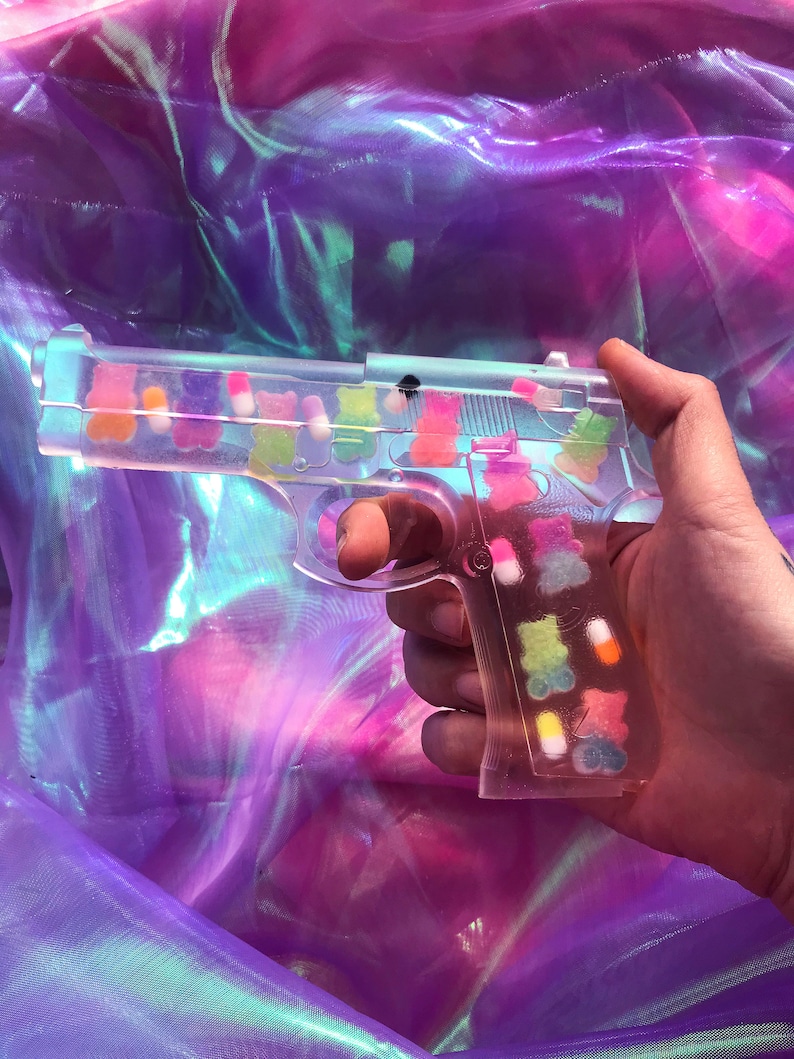 May include: A clear plastic toy gun filled with colorful gummy bears. The gun is held in a hand against a purple iridescent background.