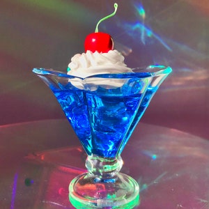 K&ouml;nnte beinhalten: Eine blaue Glasschale in Form einer Eist&uuml;te mit einer wei&szlig;en Schlagsahne-F&uuml;llung und einer roten Kirsche obenauf. Die Schale steht auf einer reflektierenden Oberfl&auml;che.