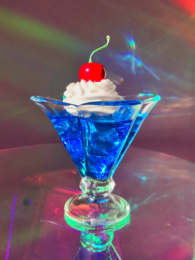 K&ouml;nnte beinhalten: Eine blaue Glasschale in Form einer Eist&uuml;te mit einer wei&szlig;en Schlagsahne-F&uuml;llung und einer roten Kirsche obenauf. Die Schale steht auf einer reflektierenden Oberfl&auml;che.