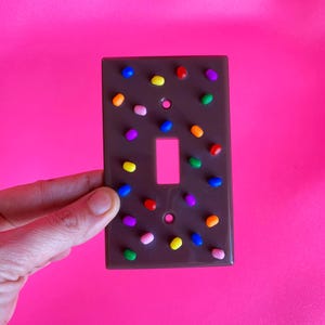 Puede incluir: Una cubierta de interruptor de luz marrón decorada con chispas coloridas y redondeadas. La cubierta tiene una abertura rectangular para el interruptor y está sobre un fondo rosa brillante.