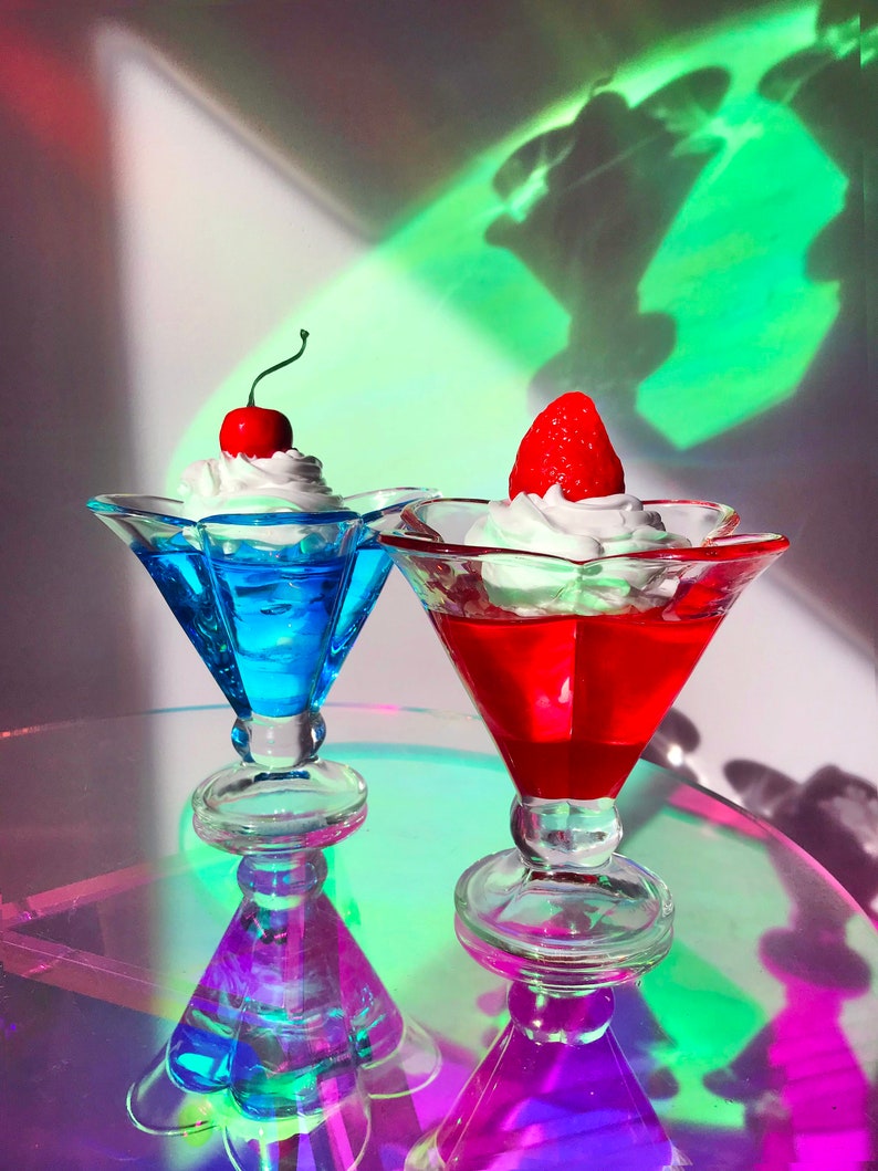 K&ouml;nnte beinhalten: Zwei klare Glasdessertschalen mit einem wei&szlig;en Schlagsahne-Topping. Eine Schale hat eine rote Kirsche oben und blaue Gelee darin. Die andere Schale hat eine rote Erdbeere oben und rote Gelee darin. Die Schalen stehen auf einer reflektierenden Oberfl&auml;che.