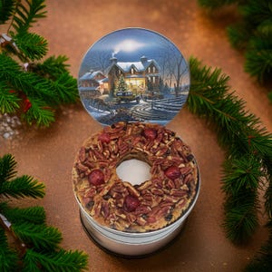 May include: A festive holiday fruitcake in a silver tin, topped with pecans and cherries. A decorative plate with a winter scene of a house with a Christmas tree is behind the cake. Green pine branches frame the scene.