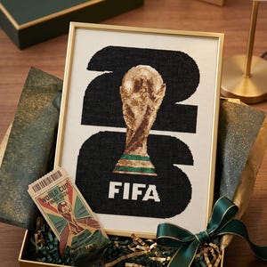 May include: A framed cross-stitch artwork featuring the FIFA World Cup trophy centered within the large black numbers 26, with the text FIFA below. Beside it sits a World Cup Final 2026 ticket, all presented in a gift box with green ribbon.