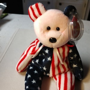 May include: A pink and white Beanie Baby bear wearing a red, white, and blue American flag outfit. The bear has a black nose and black eyes. The bear is sitting on a white surface.