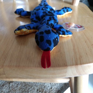 May include: A blue and black spotted plush lizard with a red tongue. The lizard is lying on a wooden surface.