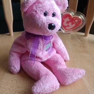 May include: Pink Beanie Baby plush toy with a purple ribbon and a Ty tag. The tag has a red heart with a white star and the words "Beanie Baby" on it.