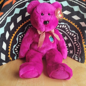 Puede incluir: Un oso de peluche rosa Beanie Baby con una cinta dorada y un parche de la Tierra verde y azul con el año 2000 en él.