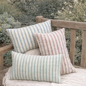 May include: Four striped throw pillows in shades of blue, red, and green rest on a wooden bench. The pillows have a vertical stripe pattern and are arranged on a cream-colored fringed blanket, creating a cozy outdoor scene.