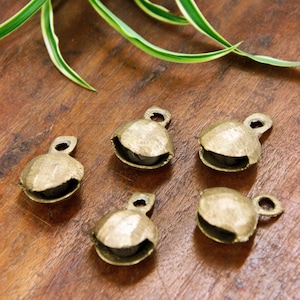 May include: Five gold-colored metal bells with a rounded shape and a small loop at the top. The bells are arranged in a semi-circle on a wooden surface.