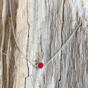 May include: A delicate silver necklace featuring a small, round, red gemstone pendant. The pendant is set in a silver bezel and hangs from a fine chain. The necklace is displayed against a rustic wooden background.