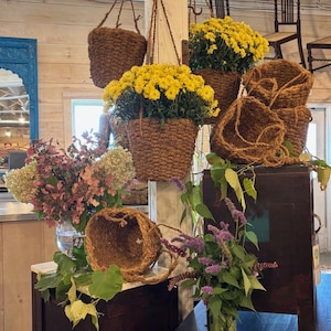 May include: A display of hanging woven baskets filled with vibrant yellow flowers. Several other empty woven baskets are also displayed. The scene includes a vase of pink and green flowers, and other greenery, creating a rustic, natural aesthetic.