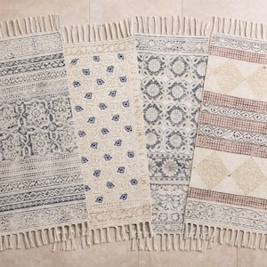 May include: Four rectangular rugs with fringed edges are displayed. Each rug features a unique pattern in shades of gray, beige, and brown. The designs include geometric shapes and floral motifs, creating a bohemian aesthetic. These rugs are suitable for home decor.