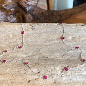 May include: A delicate silver necklace featuring small, round, vibrant red gemstones evenly spaced along the chain. The necklace is displayed on a natural wood surface, showcasing its elegant design and color contrast.
