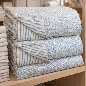 May include: A stack of three folded quilts featuring blue and white patterns. Each quilt has a quilted texture and displays a reversible design with one side showing blue floral or geometric prints and the other side featuring blue and white stripes.