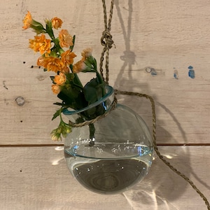 May include: A clear glass vase with a rope handle, filled with water and orange flowers. The vase is hanging from a wooden wall.