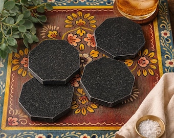 May include: Four black granite coasters with a textured surface are arranged on a decorative wooden tray. The tray features a colorful floral design with red, yellow, and blue accents. A glass of amber liquid with ice sits on a coaster.