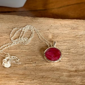 May include: A silver necklace with a round, deep red pendant. The pendant has a textured surface and is set in a silver bezel. The chain is delicate and has a small clasp. The necklace is displayed on a wooden surface.