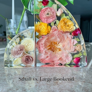 May include: Two clear resin bookends, one small and one large, each containing preserved flowers in shades of pink, yellow, and red. The text "Small vs. Large Bookend" is at the bottom. The bookends are on a reflective surface.