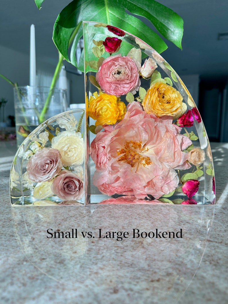 May include: Two clear resin bookends, one small and one large, each containing preserved flowers in shades of pink, yellow, and red. The text "Small vs. Large Bookend" is at the bottom. The bookends are on a reflective surface.