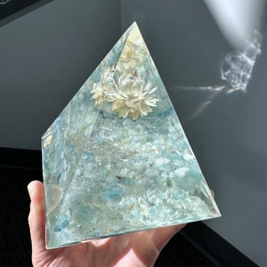 May include: A clear glass pyramid filled with light blue aquamarine stones and white dried flowers. The pyramid is held in a hand.
