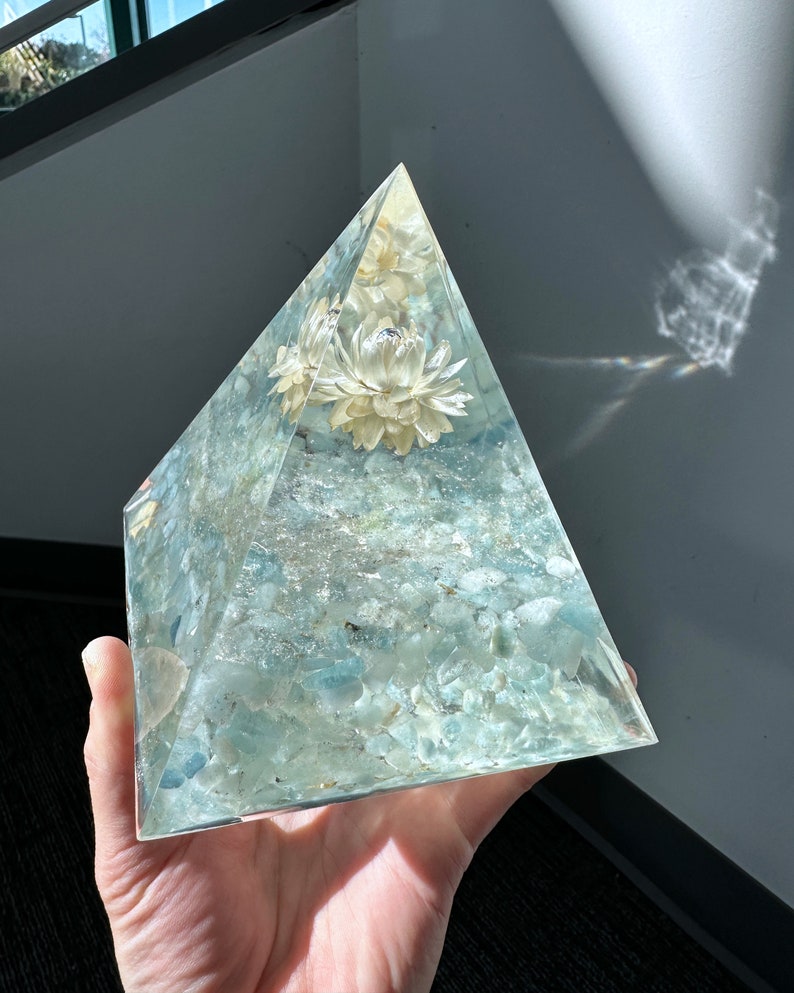 May include: A clear glass pyramid filled with light blue aquamarine stones and white dried flowers. The pyramid is held in a hand.