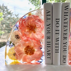 May include: A clear resin bookend with embedded coral-colored peonies, white and yellow flowers, and blue accents. The bookend is next to three white books with the phrases "ALL YOUR HEART", "DO IT WITH", and "WHATEVER YOU DO" printed on the spines.