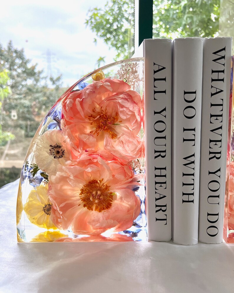 May include: A clear resin bookend with embedded coral-colored peonies, white and yellow flowers, and blue accents. The bookend is next to three white books with the phrases "ALL YOUR HEART", "DO IT WITH", and "WHATEVER YOU DO" printed on the spines.