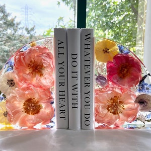 May include: Clear resin bookends with embedded flowers in shades of pink, yellow, and blue. The bookends are in a half-circle shape and hold three white books with the text "ALL YOUR HEART", "DO IT WITH", and "WHATEVER YOU DO".
