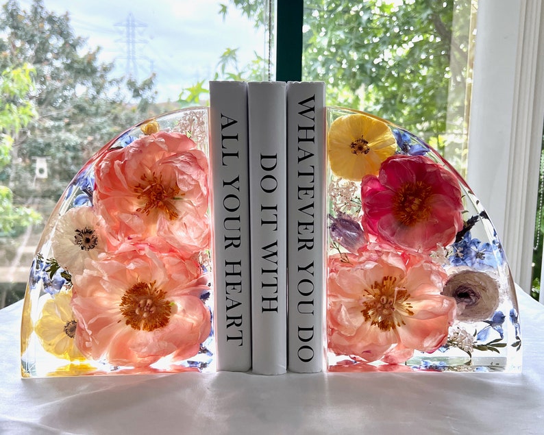 May include: Clear resin bookends with embedded flowers in shades of pink, yellow, and blue. The bookends are in a half-circle shape and hold three white books with the text "ALL YOUR HEART", "DO IT WITH", and "WHATEVER YOU DO".