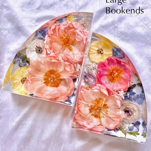 May include: Two clear resin bookends with embedded dried flowers. The bookends are semi-circular and feature peach, pink, yellow, and white flowers, along with blue accents. The text "Large Bookends" is visible in the image.
