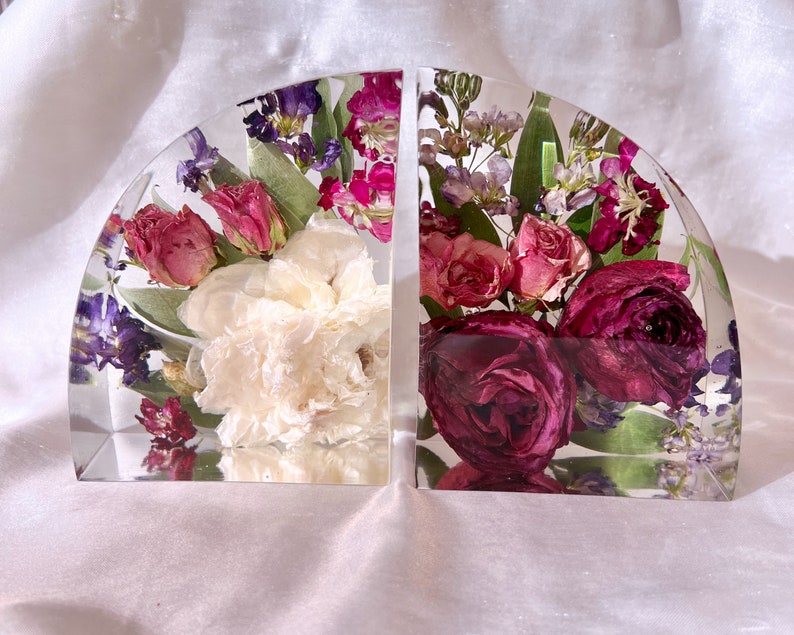 May include: Two clear, semi-circular bookends with dried flowers encased in resin. The bookends feature a variety of flowers in shades of white, pink, purple, and burgundy. The bookends are a decorative item for a bookshelf.