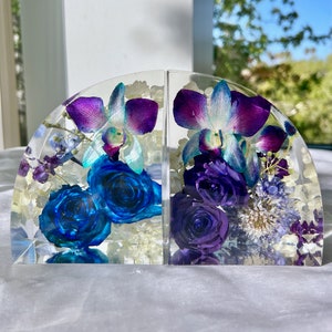 May include: Two clear resin bookends, each with a half-circle shape, showcasing vibrant blue roses and purple orchids. The flowers are encased in clear resin, creating a decorative and functional item for any bookshelf.