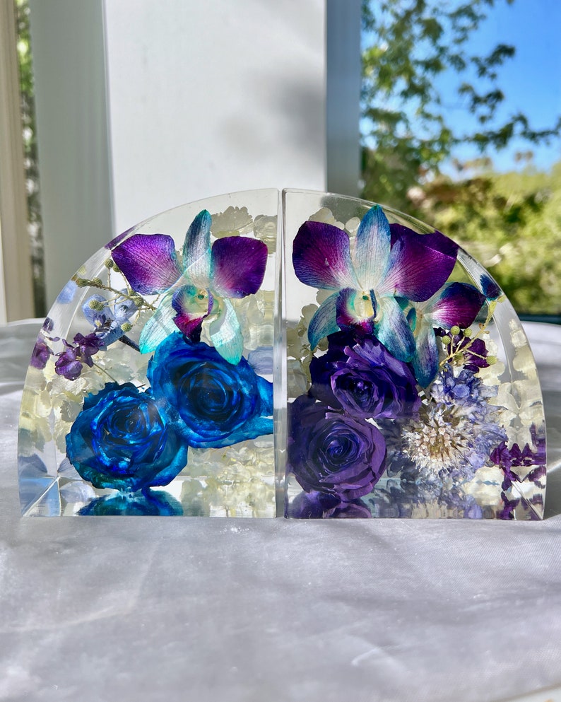 May include: Two clear resin bookends, each with a half-circle shape, showcasing vibrant blue roses and purple orchids. The flowers are encased in clear resin, creating a decorative and functional item for any bookshelf.