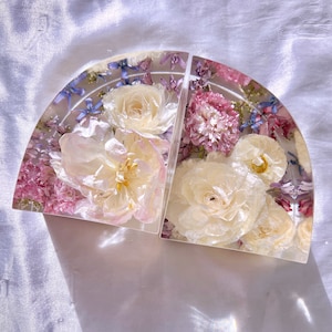 May include: Two clear, semi-circular bookends with embedded dried flowers. The bookends feature a variety of flowers in shades of white, pink, and purple, encased in clear resin. The bookends are arranged side by side.