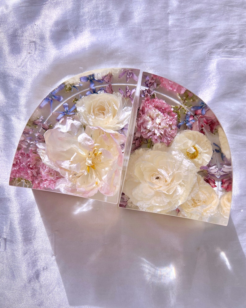 May include: Two clear, semi-circular bookends with embedded dried flowers. The bookends feature a variety of flowers in shades of white, pink, and purple, encased in clear resin. The bookends are arranged side by side.
