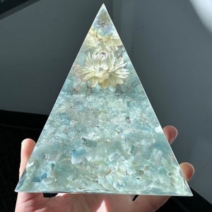 May include: A clear glass pyramid filled with aquamarine stones and a dried white flower. The pyramid is held in a hand. Aquamarine Stones.