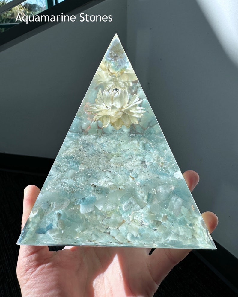 May include: A clear glass pyramid filled with aquamarine stones and a dried white flower. The pyramid is held in a hand. Aquamarine Stones.