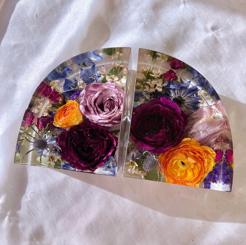 May include: Two clear resin bookends, each in a semi-circular shape, encasing dried flowers. The flowers include purple, orange, and blue blooms. The bookends are set against a white, textured background.