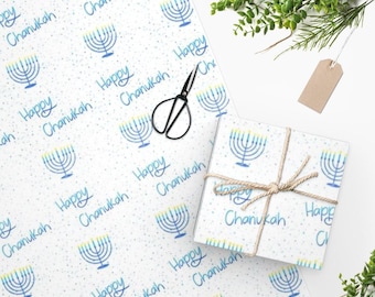 Puede incluir: Papel de regalo blanco con un diseño de menorá azul y las palabras "Happy Chanukah". También se ven una caja de regalo envuelta, unas tijeras y una etiqueta en blanco.